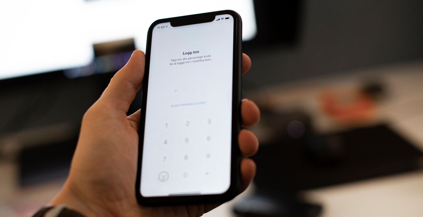 Mann som holder telefon med bank-app foran PC-skjerm