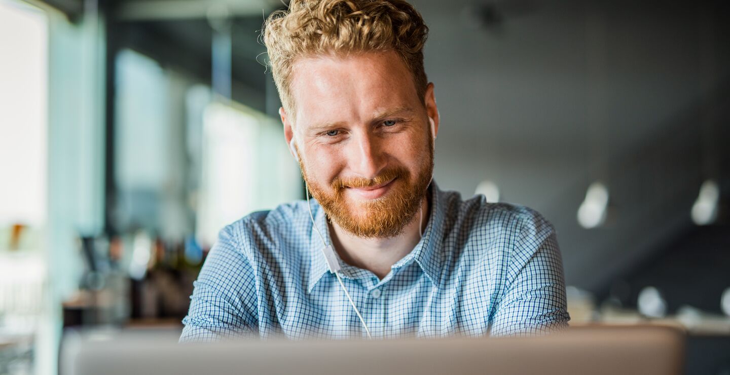 Smilende mann med ørepropper foran en mac. (Foto: Djelics).