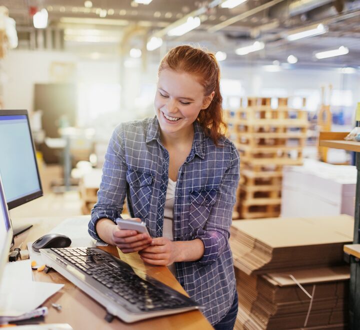 Kvinne med mobil ved pult og PC