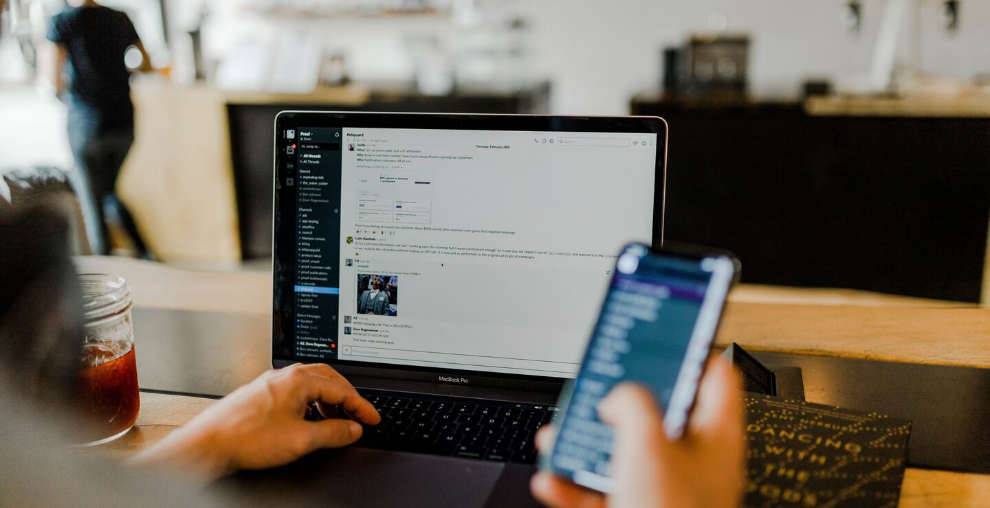 En person som sitter foran PC'en og jobber mens han holder i en mobiltelefon.