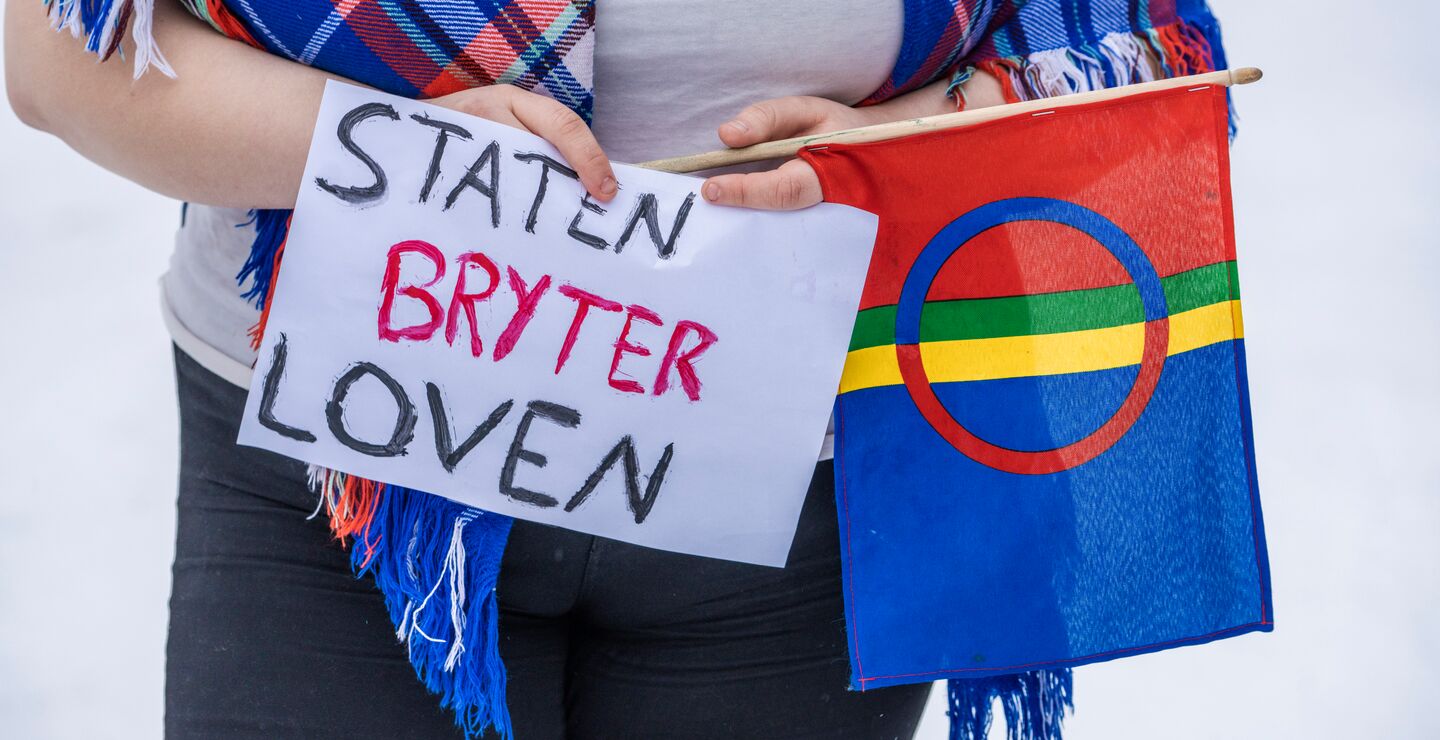 Ung kvinne holder samisk flagg og plakat der det står 'Staten bryter loven'