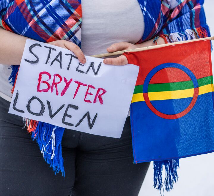 Ung kvinne holder samisk flagg og plakat der det står 'Staten bryter loven'