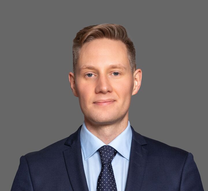 Professional headshot of a Daniel Kinn wearing a dark blue suit, light blue shirt, and a navy tie with white polka dots. He has short, neatly styled blond hair and a neutral expression, standing against a plain grey background