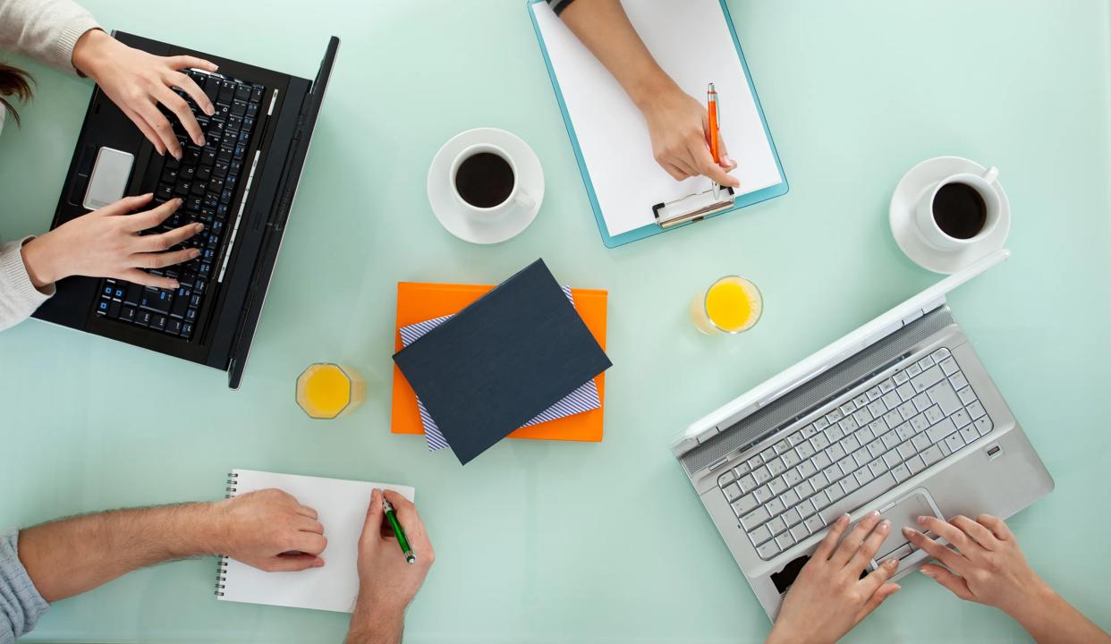 Four people working around a table. The image shows their hands, each working on either a PC or writing notes in a book.