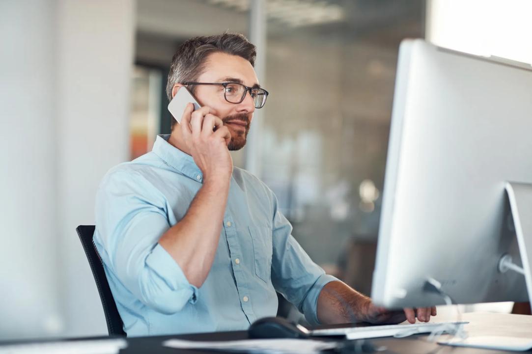 Bilde av en forretningsmann som bruker en datamaskin og mobiltelefon ved skrivebordet sitt på et moderne kontor.