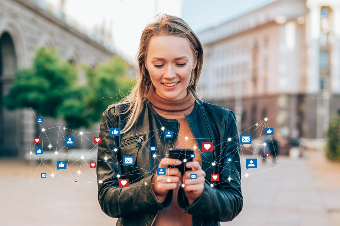 Kvinne står på en plass og holder mobiltelefon.  Rundt telefonen vises likes og emoticons