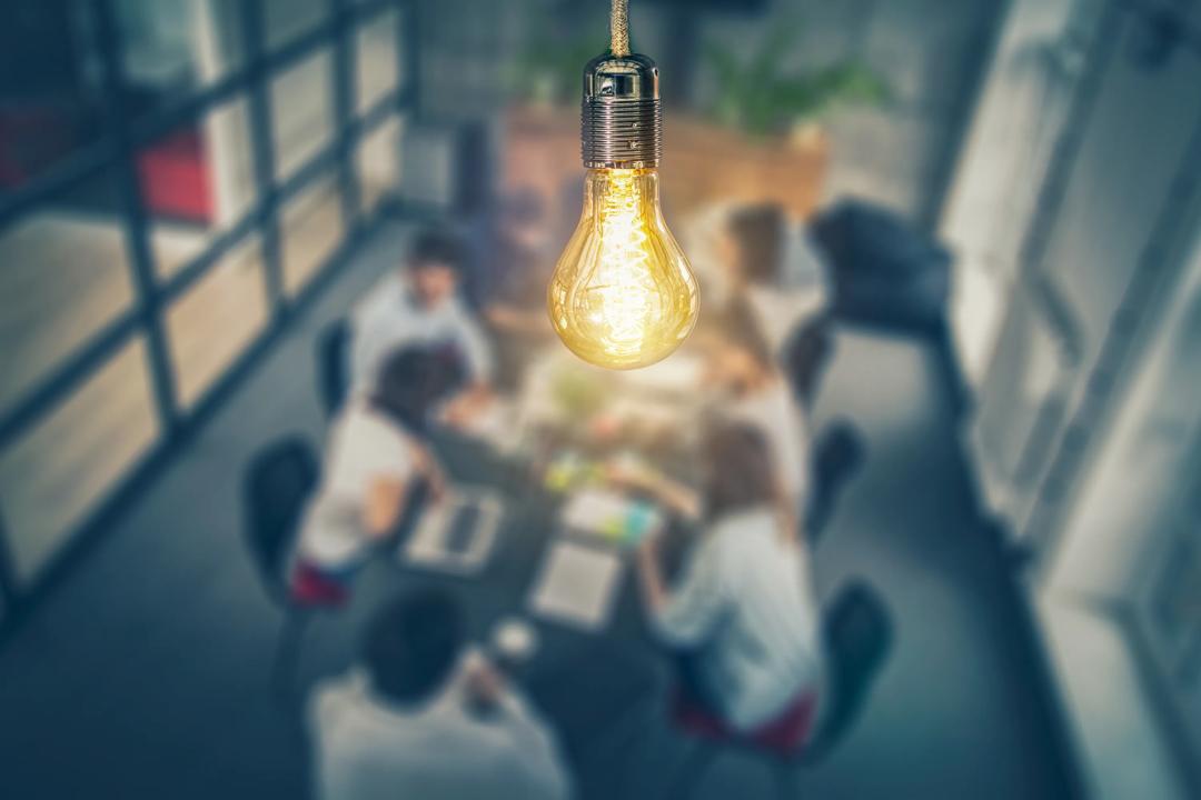 A group of people working together around a table. A light bulb hanging from the roof and is in focus in the image.