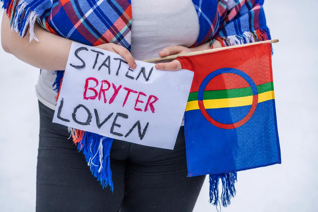 Ung kvinne holder samisk flagg og plakat der det står 'Staten bryter loven'