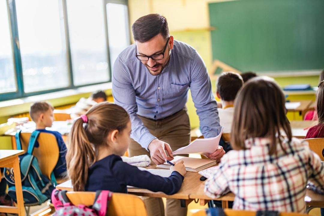 En barneskolelærer som står og hjelper to elever.