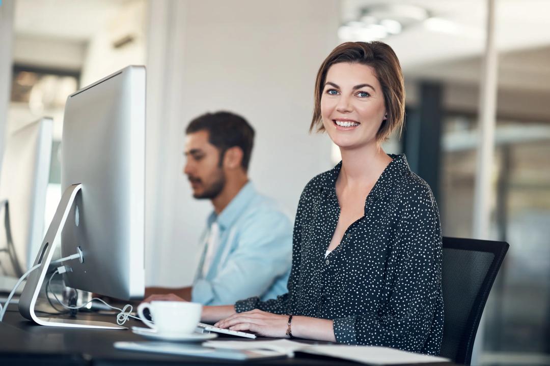 Bilde av en kvinne og en mann som jobber foran hver sin PC. Damen smiler til kamera.