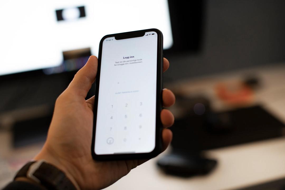 Mann som holder telefon med bank-app foran PC-skjerm