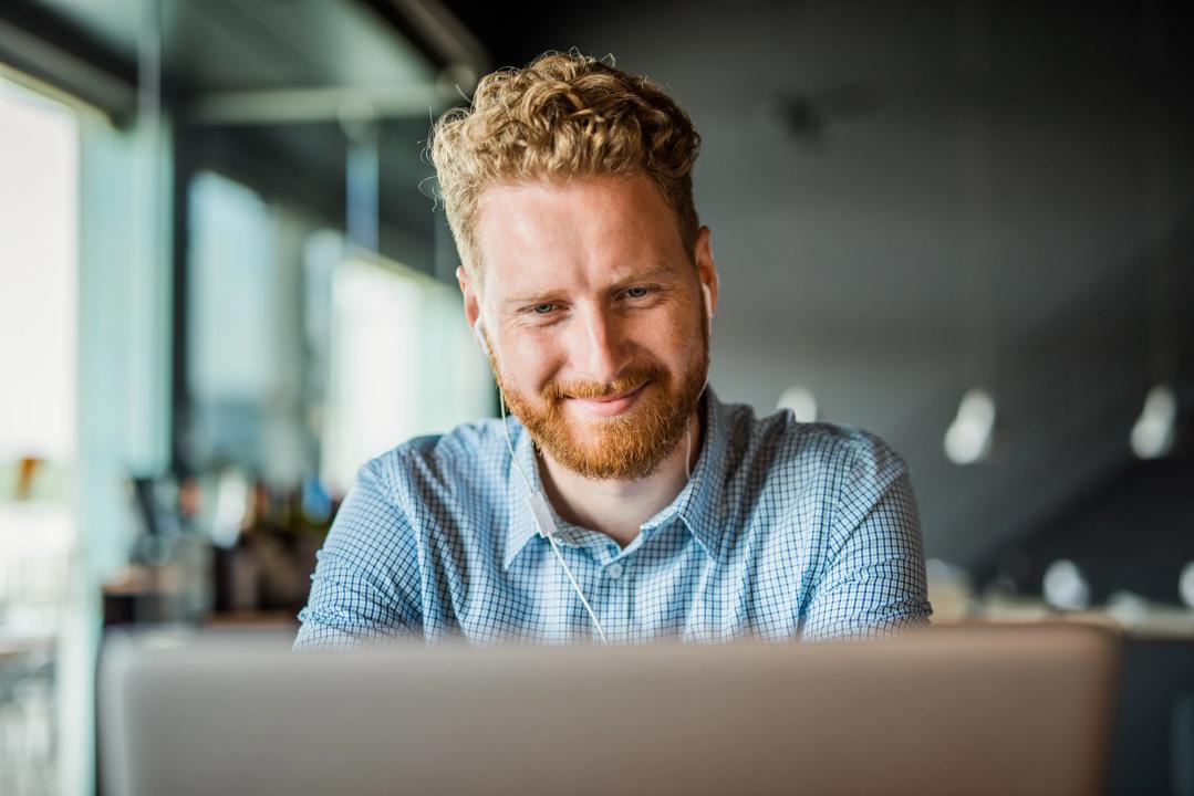 Smilende mann med ørepropper foran en mac. (Foto: Djelics).