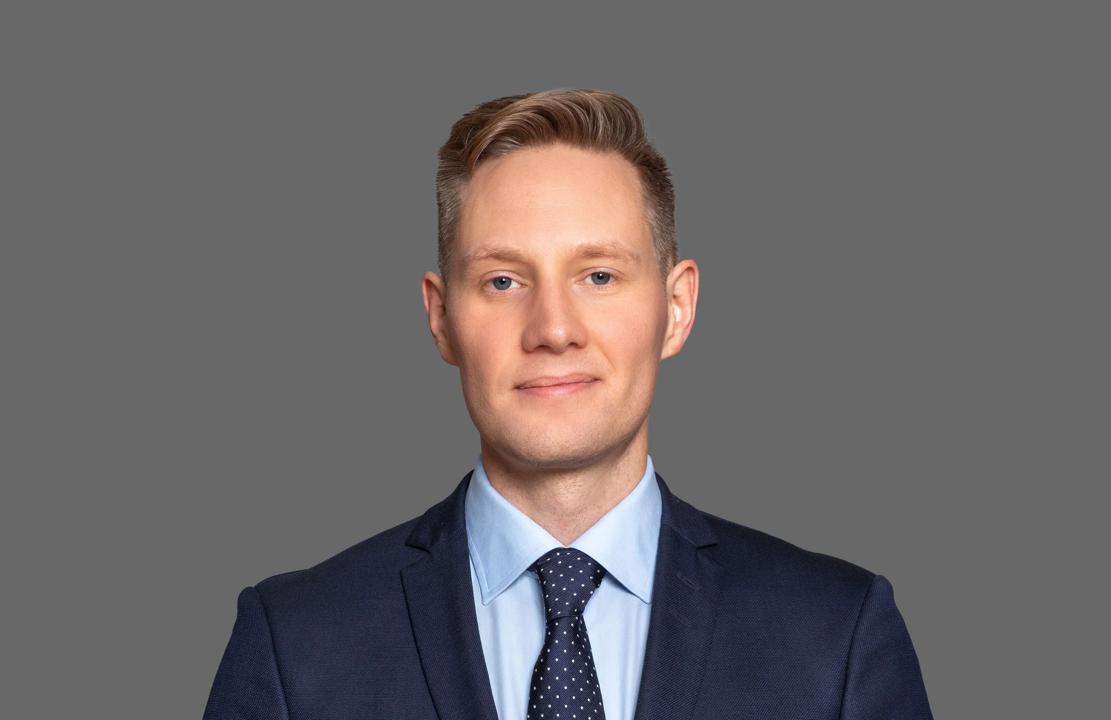 Professional headshot of a Daniel Kinn wearing a dark blue suit, light blue shirt, and a navy tie with white polka dots. He has short, neatly styled blond hair and a neutral expression, standing against a plain grey background